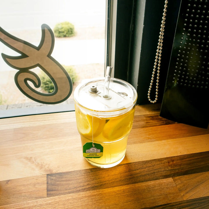 Delikatesi i tako to... Ahmad Tea lemon green tea served in a glass cup with a lid and straw on a wooden table