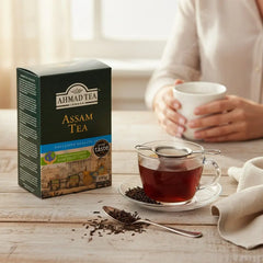 Delikatesi i tako to... Ahmad Tea Assam loose leaf tea box with a steaming cup of tea and tea leaves on a table.