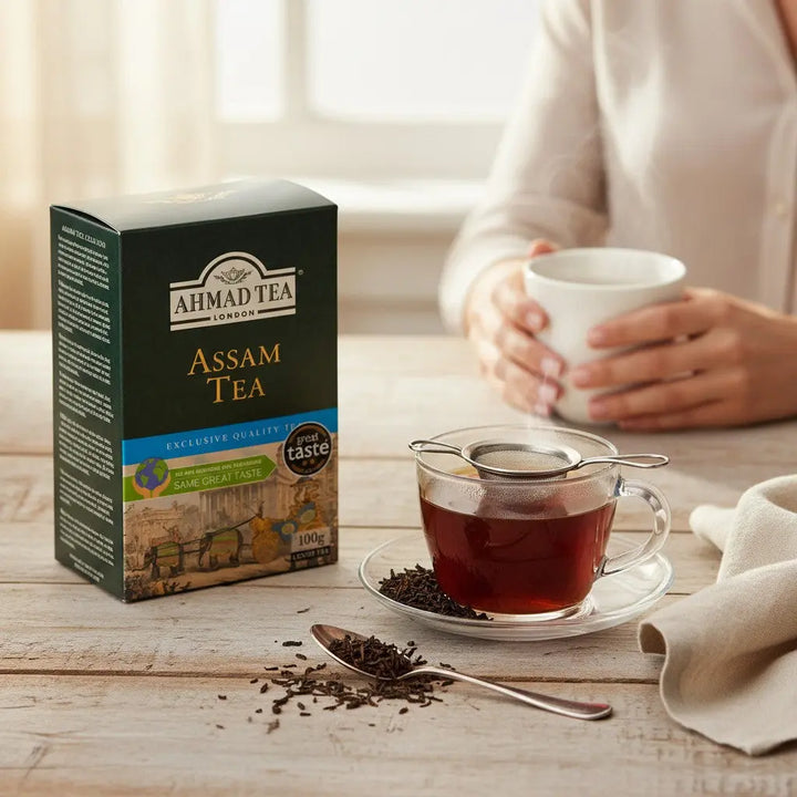 Delikatesi i tako to... Ahmad Tea Assam loose leaf tea box with a steaming cup of tea and tea leaves on a table.