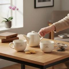 Delikatesi i tako to... white porcelain Ahmad Tea teapot and two mugs on a wooden table with cookies and infuser